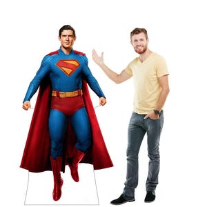 May include: A life-size cardboard cutout of Superman in his iconic blue suit with red trunks, boots, and cape. The Superman logo is prominently displayed on the chest. A person in a yellow shirt and jeans stands next to the cutout.