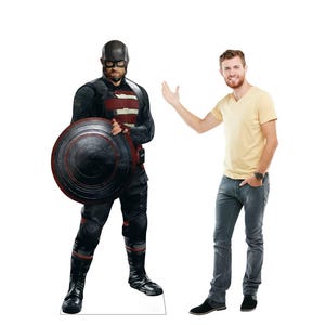 May include: A life-size cardboard cutout of a character in a black and red costume, holding a shield. The character wears a mask and boots. A person in a yellow shirt and jeans stands next to the cutout, gesturing with their hand.