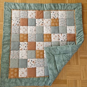 Könnte beinhalten: Eine grüne und braune Patchwork-Decke mit Blumenmuster. Die Decke ist in der Mitte gefaltet und hat einen grünen Rand.