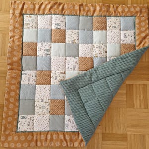 Könnte beinhalten: Eine Patchwork-Babydecke mit einem braunen und weißen Blumenrand. Die Decke besteht aus verschiedenen Stoffen mit unterschiedlichen Mustern, darunter Tiere, Blumen und geometrische Formen. Die Decke ist in der Hälfte gefaltet und zeigt auf der Rückseite einen einfarbigen grünen Stoff.