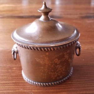 May include: A small, round, copper container with a lid and two decorative handles. The container has a floral design etched into the side.
