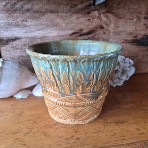 Puede incluir: Una maceta de cerámica hecha a mano con un diseño texturizado. La maceta presenta un interior turquesa y un exterior marrón con patrones tallados de hojas y tejido de cesta. El borde es de un marrón más oscuro y la pieza tiene una estética rústica y artesanal.