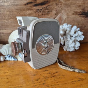 May include: A vintage 8mm film camera with a brown and beige body, a silver lens, and a black strap. The camera has a silver dial with the text "8mm" on it.
