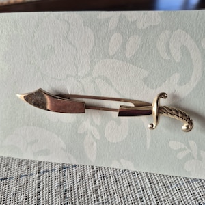 Puede incluir: Un broche dorado con forma de espada pequeña. El broche tiene un cierre tipo imperdible. El mango de la espada está detallado con un diseño de cuerda trenzada. La hoja es curva y tiene una punta roma.
