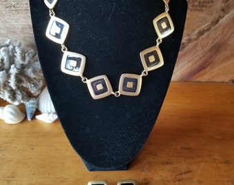 May include: A gold necklace with black square-shaped pendants. The necklace is on a black velvet display stand. There are two matching earrings with black square-shaped pendants.