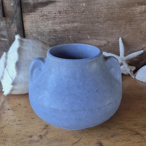 May include: A small, round, blue ceramic vase with two handles. The vase has a matte finish and is sitting on a wooden surface. A white seashell and starfish are in the background, suggesting a coastal theme.