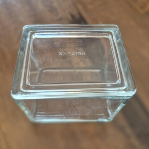 Puede incluir: Tarro cuadrado de vidrio transparente con tapa rectangular. La palabra "WHEATON" está grabada en la tapa. El tarro está sobre una superficie de madera.