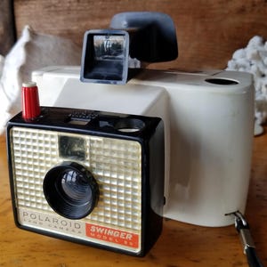 May include: A white Polaroid Swinger Model 30 Land Camera with a black lens and viewfinder. The camera has a red button on the side and a black strap.
