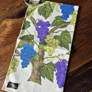 May include: A rectangular linen towel with a grape vine design in shades of purple, blue, green, and brown. The towel has a white background and a tag that reads "Kay Pee". A magnifying glass rests on the towel.
