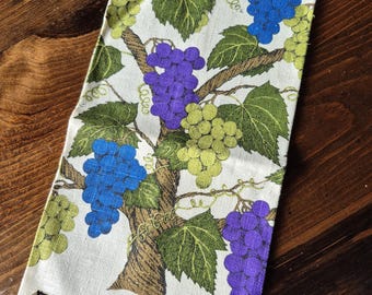 May include: A rectangular off-white linen towel featuring a printed pattern of grapevines with green leaves, brown branches, and clusters of blue, purple, and light green grapes. A small black fabric tag reads KarDee 100% Pure Linen.