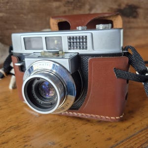 May include: A vintage silver and black Tomatic 35mm camera with a brown leather case. The camera lens is prominent, with the words "Tomatic 35 Camera" inscribed on the lens. The camera strap is black and white.