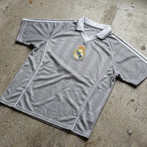 May include: Gray short-sleeve soccer jersey with white accents and a white collar. The jersey features a team emblem on the chest and white stripes on the shoulders. The shirt has a vertical striped pattern.