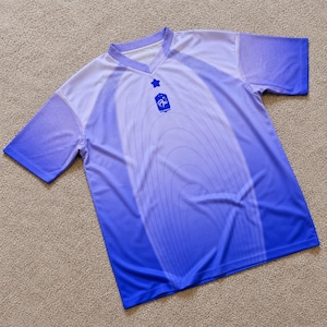 May include: A short-sleeve V-neck jersey featuring a blue and white gradient design. The chest displays a small blue star above a shield-shaped emblem containing a rooster graphic. The front is decorated with subtle, concentric oval line patterns.