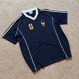 Pu&ograve; includere: Una maglia a maniche corte blu navy con colletto a V bianco, profili bianchi lungo i lati e strisce bianche sulle spalle. Il petto presenta un emblema dorato a forma di gallo con due stelle sopra e un logo KDN dorato sul lato destro.