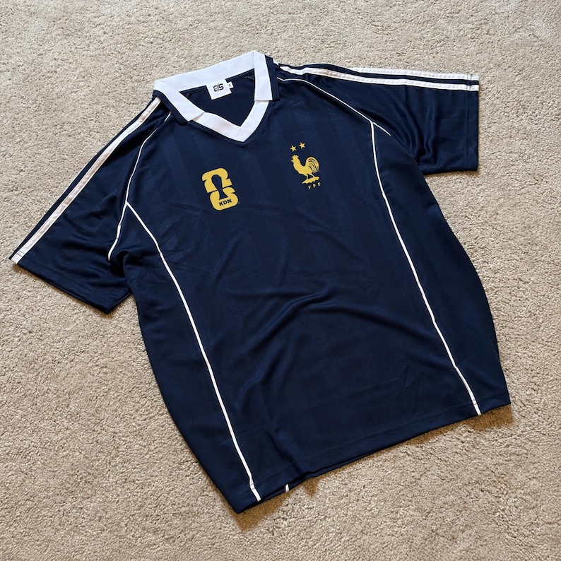 Pu&ograve; includere: Una maglia a maniche corte blu navy con colletto a V bianco, profili bianchi lungo i lati e strisce bianche sulle spalle. Il petto presenta un emblema dorato a forma di gallo con due stelle sopra e un logo KDN dorato sul lato destro.