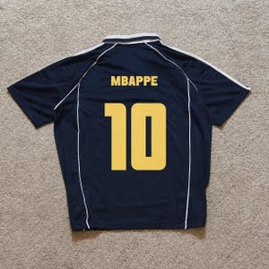 Pu&ograve; includere: Una maglia sportiva a maniche corte blu navy scuro con colletto bianco e sottili profili bianchi lungo le spalle e i fianchi. Sul retro &egrave; riportato il nome MBAPPE in lettere maiuscole gialle sopra il grande numero 10 giallo.