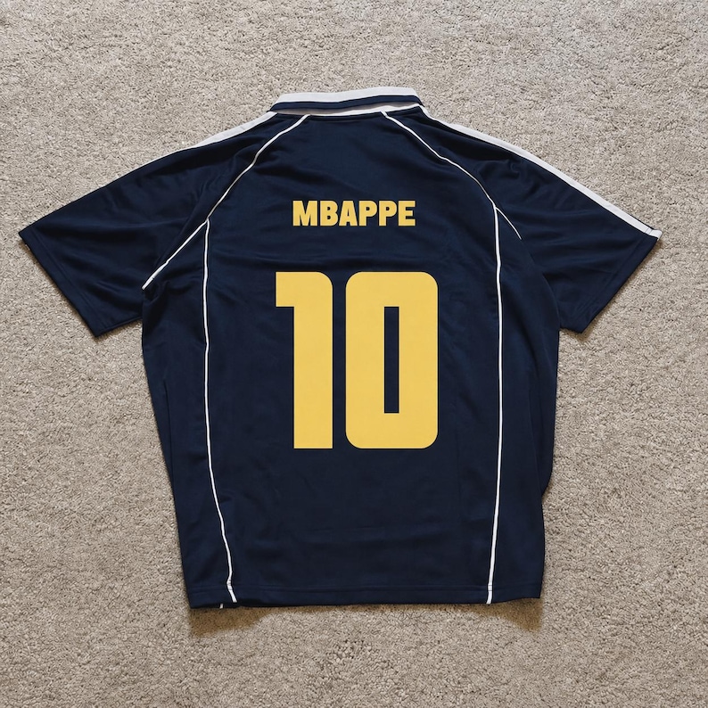 Pu&ograve; includere: Una maglia sportiva a maniche corte blu navy scuro con colletto bianco e sottili profili bianchi lungo le spalle e i fianchi. Sul retro &egrave; riportato il nome MBAPPE in lettere maiuscole gialle sopra il grande numero 10 giallo.
