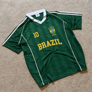 Puede incluir: Camiseta de fútbol verde de Brasil con detalles blancos. La camiseta muestra el número 10, la palabra "BRAZIL" en amarillo y un emblema de la bandera de Brasil. Tiene cuello en V y rayas blancas en las mangas.