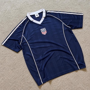 Pode incluir: Uma camisola de manga curta azul-marinho com gola branca e detalhes em vivo branco. A peça apresenta um padrão subtil de riscas verticais e um emblema central em forma de escudo com o texto USA, riscas vermelhas e brancas e estrelas azuis.