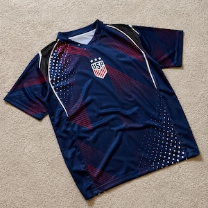 May include: A navy blue short-sleeve athletic jersey featuring a V-neck collar and a central USA crest logo with three stars. The shirt displays a graphic pattern of thin red diagonal lines and white dots across the front, with black shoulder panels.