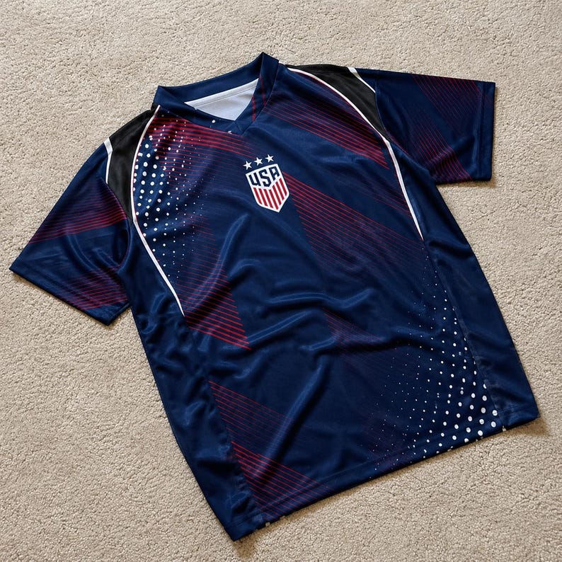 May include: A navy blue short-sleeve athletic jersey featuring a V-neck collar and a central USA crest logo with three stars. The shirt displays a graphic pattern of thin red diagonal lines and white dots across the front, with black shoulder panels.