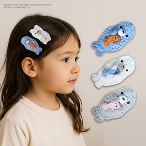 May include: Four crocheted seal hair clips. Each clip features a seal holding a fish in its mouth. The seals are in different colours: blue, white, and grey.