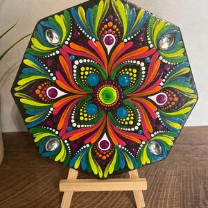 Puede incluir: Diseño de mandala pintado a mano y colorido sobre una placa de madera octogonal negra. La obra de arte presenta tonos vibrantes de naranja, rosa, verde, azul y blanco, acentuados con pequeños adornos brillantes. El mandala se exhibe sobre un pequeño caballete de madera.