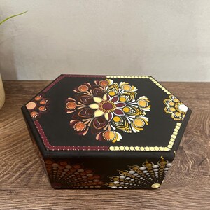 May include: A black, hexagonal wooden box with a detailed mandala design on the lid. The mandala features a floral pattern in shades of yellow, orange, and burgundy. The box is decorated with dot art.