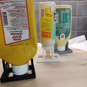 Könnte beinhalten: Drei Gewürzflaschen in Haltern auf einer Holzoberfläche. Die Flaschen sind gelb und weiß, mit Etiketten. Eine Flasche ist mit "Johnny's" und "SÖTSTARK SENAP" beschriftet. Die andere Flasche ist mit "GE HELGEN EXTRA ALLT!" beschriftet.