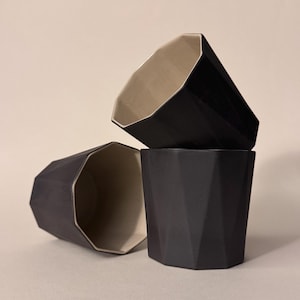 Puede incluir: Tres tazas geométricas negras de cerámica están apiladas. Las tazas tienen un diseño facetado y un interior beige claro. Las tazas están dispuestas sobre un fondo neutro, mostrando su diseño moderno y colores contrastantes.
