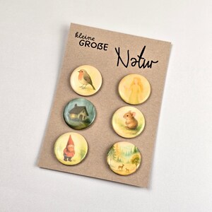 Könnte beinhalten: Set aus sechs runden Knöpfen mit Naturmotiven, darunter Illustrationen eines Vogels, Hauses, Zwergs, Engels, einer Maus und einer Waldszene. Die Knöpfe sind auf einer braunen Karte mit den Worten "kleine GROBE Natur" in schwarzer Schrift angeordnet.