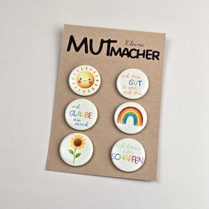 Könnte beinhalten: Ein Set aus sechs weißen Ansteckbuttons mit farbenfrohen Designs und deutschem Text, darunter eine lächelnde Sonne, ein Regenbogen und eine Sonnenblume. Die Buttons sind auf einer braunen Karte mit dem Wort "MUTMACHER" in Schwarz angeordnet.