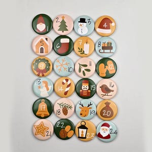Könnte beinhalten: Ein Set aus 24 runden Knöpfen mit Weihnachtsmotiven, jeweils mit einer einzigartigen Illustration und einer Nummer von 1 bis 24. Designs umfassen einen Zwerg, einen Weihnachtsbaum, einen Schneemann, einen Weihnachtsmannhut, ein Haus, einen Strumpf, eine Kerze, Geschenke, eine Schleife, einen Schneeflocke, Blätter, Fäustlinge, einen Engel, eine Zuckerstange, einen Nussknacker, einen Vogel, eine Glocke, einen Tannenzapfen, ein Rentier, eine Goldtasche, einen Stern, Nüsse, eine Laterne und den Weihnachtsmann.