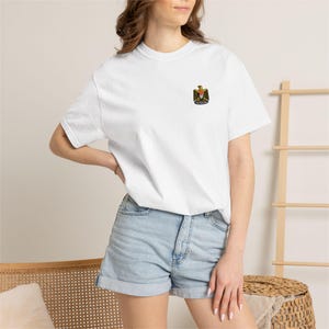 Könnte beinhalten: Ein weißes kurzärmeliges T-Shirt mit einer kleinen gestickten Grafik auf der linken Brust. Das Design zeigt einen Zwerg mit gelbem Hut, rotem Bart und schwarzem Mantel. Das T-Shirt hat einen Rundhalsausschnitt und eine normale Passform aus Baumwolle.