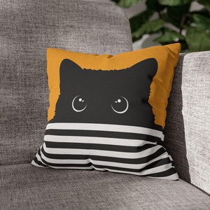 Könnte beinhalten: Ein quadratisches Dekokissen mit einer Grafik der Silhouette einer schwarzen Katze mit großen weißen Augen, die über horizontalen schwarzen und weißen Streifen hervorschaut, vor einem einfarbigen senfgelben Hintergrund im oberen Bereich.