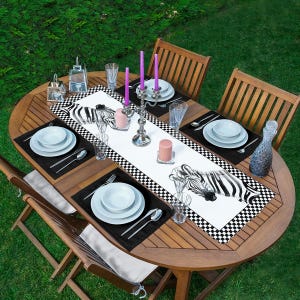 Könnte beinhalten: Ein Esstisch im Freien für sechs Personen. Der Holztisch ist mit weißen Tellern, Besteck und schwarzen Tischsets gedeckt. Ein Tischläufer mit Zebrastreifen und einem schwarz-weißen Schachbrettmuster verläuft in der Mitte. Lila Kerzen und eine dekorative Glasflasche vervollständigen die Szene.