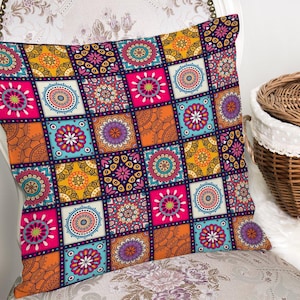 Könnte beinhalten: Quadratisches Zierkissen mit Patchwork-Design. Das Kissen zeigt ein lebendiges Muster aus Quadraten in den Farbtönen Rosa, Orange, Blau, Gelb und Lila. Jedes Quadrat enthält ein Blumen- oder Mandala-ähnliches Design. Das Kissen liegt auf einem gemusterten Stuhl.