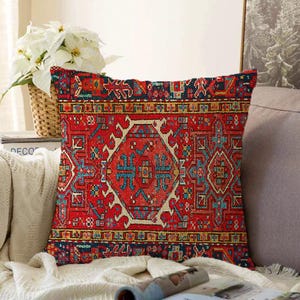 Könnte beinhalten: Ein dekoratives quadratisches Kissen mit einem aufwendigen, teppichartigen Muster. Das Kissen hat einen überwiegend roten Hintergrund mit detaillierten geometrischen Designs in Blau, Gold und anderen Farben. Das Kissen liegt auf einer weißen Decke.