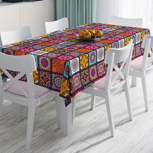 Könnte beinhalten: Ein rechteckiges Tischtuch mit einem lebendigen Patchwork-Design in Orange, Rosa, Blau und Lila bedeckt einen Esstisch. Weiße Stühle mit rosa Sitzkissen umgeben den Tisch. Ein Teller mit Orangen steht auf dem Tisch.