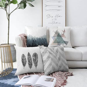 Könnte beinhalten: Eine Sammlung dekorativer Kissen mit verschiedenen Designs. Ein Kissen zeigt eine Waldszene in Blau- und Grautönen. Ein anderes zeigt einen geometrischen Hirschkopf. Weitere Kissen haben Feder- und Fischgrätmuster.