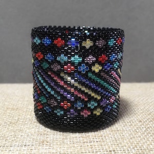 Könnte beinhalten: Ein schwarzes Perlenarmband mit einem farbenfrohen geometrischen Muster. Das Armband ist aus kleinen, bunten Perlen in den Farben Rot, Blau, Grün, Gelb und Lila gefertigt.