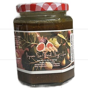 Può includere: Un barattolo di marmellata di fichi secchi al sole con coperchio a scacchi rossi e bianchi. L'etichetta presenta illustrazioni di fichi e il testo "Organic Sun-Dried Fig Jam." Peso netto 327g.