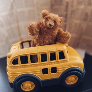 Könnte beinhalten: Ein kleiner, brauner Teddybär sitzt in einem gelben Green Toys Schulbus. Der Bus hat schwarze Räder und den Text "Green Toys School Bus" auf der Seite. Der Bär hat die Arme erhoben.
