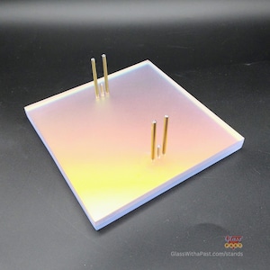Puede incluir: Un expositor cuadrado de vidrio iridiscente con tres postes de metal dorado. El expositor tiene un degradado de colores, que va del amarillo al rosa. La base tiene una forma rectangular sólida. La dirección del sitio web "GlassWithaPast.com/stands" es visible.