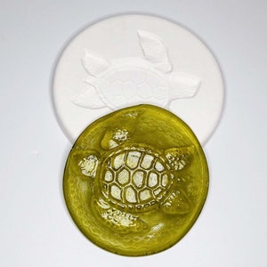 May include: A white, circular mold with a raised turtle design, next to a gold-colored, circular turtle-shaped object. The turtle design is detailed with visible shell patterns and flippers. The objects are likely for crafting or decorative purposes.
