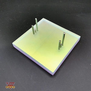 Puede incluir: Un objeto cuadrado de vidrio iridiscente con cuatro varillas metálicas de diferentes alturas. El vidrio tiene una apariencia en capas con una parte superior amarillo-verdosa y una base rosa-púrpura. El logotipo "Glass PAST" está en la esquina inferior izquierda.