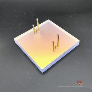 Peut inclure: Un présentoir carré en verre dépoli avec quatre poteaux en laiton de hauteurs variables. Le présentoir présente un dégradé de couleurs, passant du jaune au rose. L'adresse du site Web "GlassWithaPast.com/stands" est visible en bas à droite.