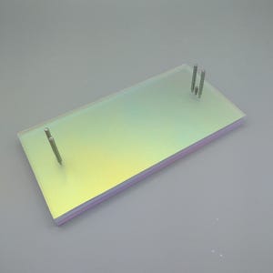 Puede incluir: Un expositor rectangular de acrílico iridiscente con tres postes metálicos. El expositor tiene un sutil degradado de color de morado a amarillo.