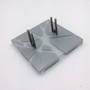 May include: A clear, frosted glass square with a raised, geometric design. Five silver metal rods are inserted into the glass.