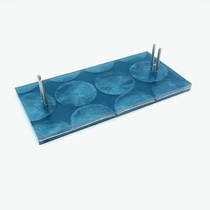 May include: A rectangular, teal-colored tray with a pattern of large, overlapping circles. The tray has three silver metal posts. The tray is made of a clear material with a teal-colored design.